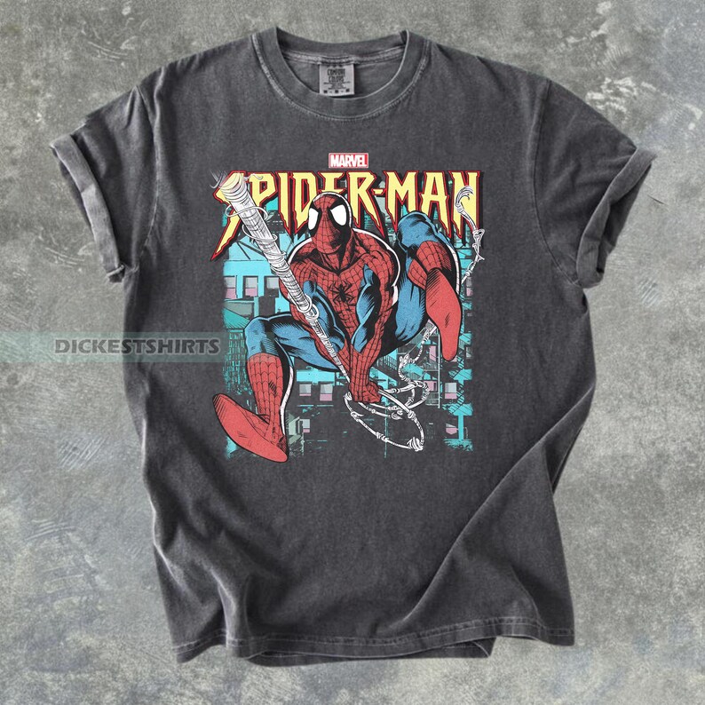 May include: Dark gray t-shirt featuring a graphic of Spider-Man in a dynamic pose, swinging from a web. The design includes the Marvel logo and the words "Spider-Man" in a bold, colorful font. The shirt has rolled-up sleeves.