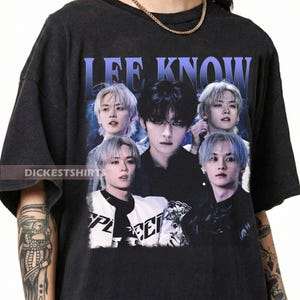 May include: Dark gray t-shirt featuring a graphic of a person with multiple images of themselves, with the text "LEE KNOW" in blue. The shirt has a vintage style and the text "DICKESTSHIRT" is printed on the bottom left.