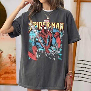 May include: Dark gray t-shirt featuring a colorful Spider-Man graphic. The design includes the Marvel logo and the words "SPIDER-MAN" in yellow and red. The superhero is depicted in his classic red and blue suit, with a web-slinging pose.