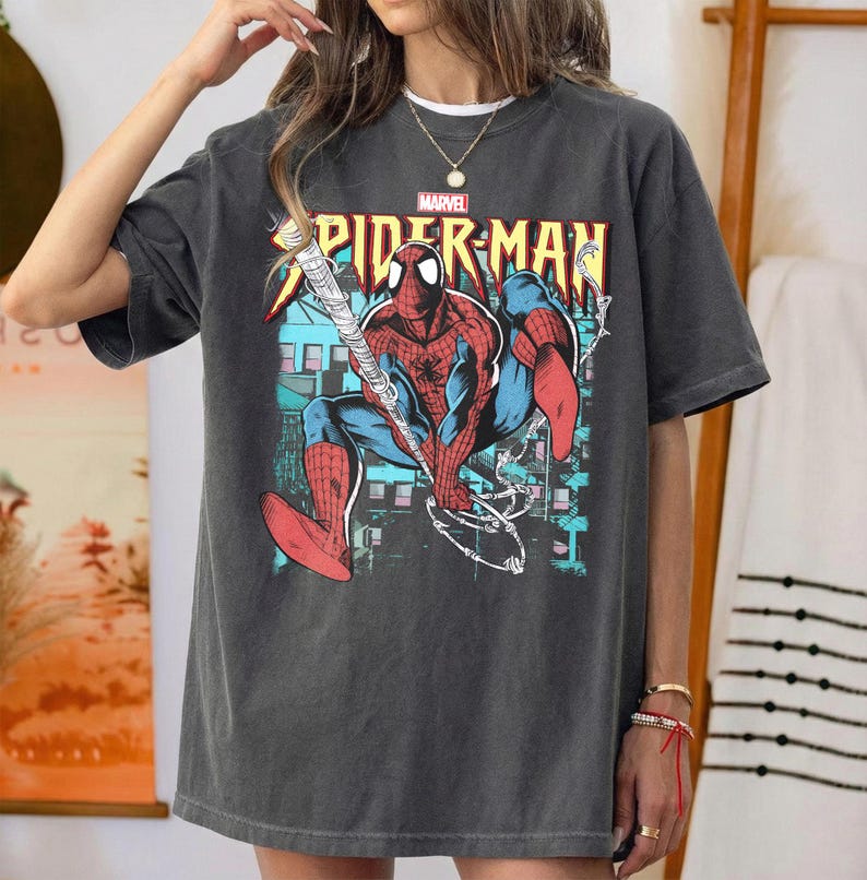 May include: Dark gray t-shirt featuring a colorful Spider-Man graphic. The design includes the Marvel logo and the words "SPIDER-MAN" in yellow and red. The superhero is depicted in his classic red and blue suit, with a web-slinging pose.