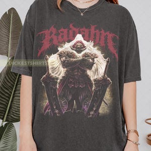 May include: Dark gray t-shirt featuring a detailed illustration of a warrior with a long white beard, holding two large swords. The word "Radahn" is printed in red above the warrior. The shirt has a vintage, worn look.