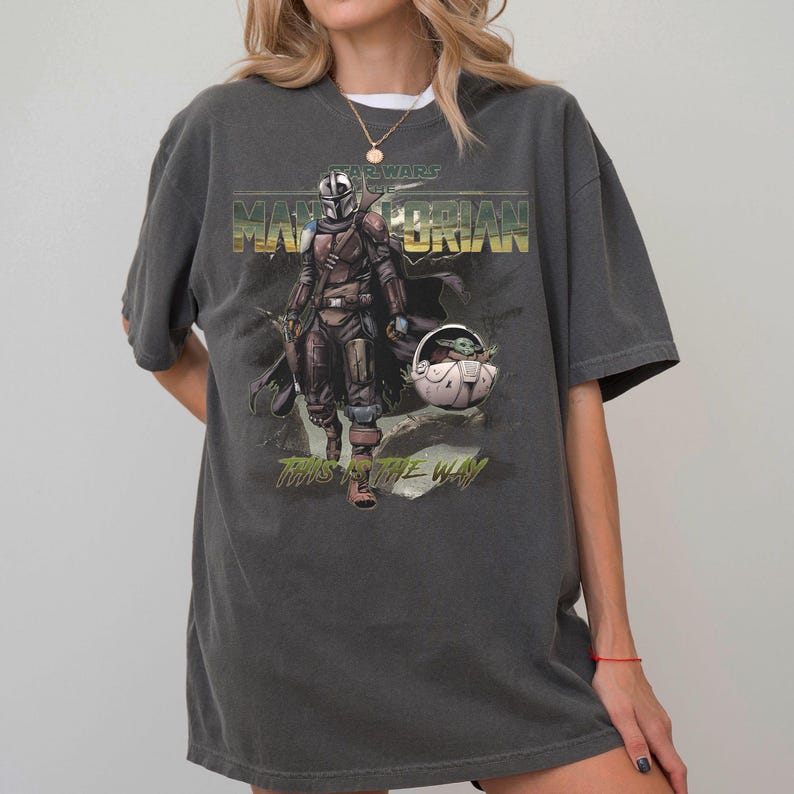 May include: Dark gray t-shirt featuring a graphic of The Mandalorian and Grogu from Star Wars. The design includes the text "STAR WARS" above "THE MANDALORIAN" in gold lettering, and "THIS IS THE WAY" below the image. The shirt has short sleeves.
