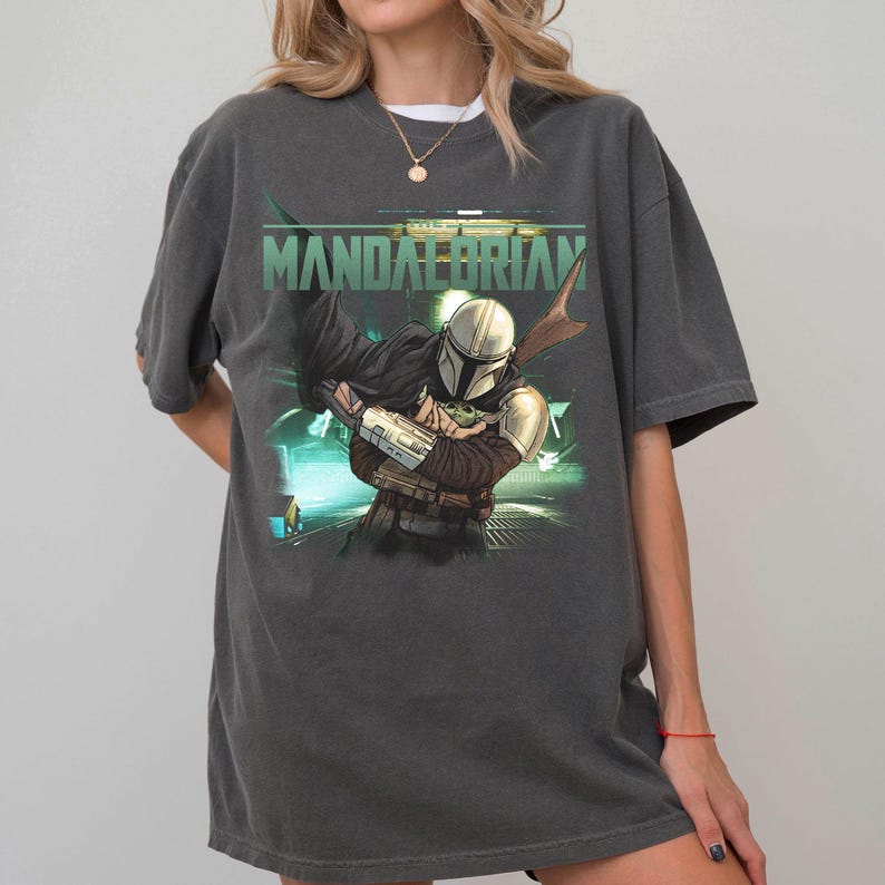 May include: Dark gray t-shirt featuring a graphic of The Mandalorian and Grogu. The design includes the text "MANDALORIAN" in green above the illustration. The artwork depicts the Mandalorian in armor, holding Grogu, with a futuristic background.