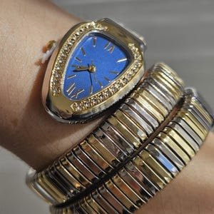 Può includere: Un orologio da polso color oro e argento con quadrante blu e numeri romani dorati. L'orologio ha un quadrante triangolare con un bordo a forma di diamante. Il cinturino è un design flessibile in oro e argento.