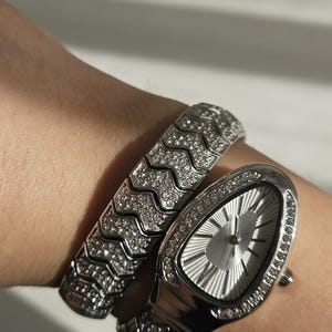 Puede incluir: Un conjunto de reloj y pulsera plateados. El reloj tiene una esfera ovalada plateada y está adornado con pequeñas piedras brillantes. La pulsera a juego presenta un diseño en zigzag y también está engastada con piedras.