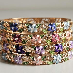 Può includere: Una pila di braccialetti dorati con motivi floreali. Ogni braccialetto presenta fiori smaltati colorati in tonalità di blu, rosa, viola e verde, accentuati da piccoli cristalli. I braccialetti hanno un design traforato.