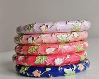 May include: A stack of five enamel bangles in shades of pink, red, and blue. Each bangle is adorned with a floral design featuring white flowers and green leaves. The bangles are stacked on top of each other.