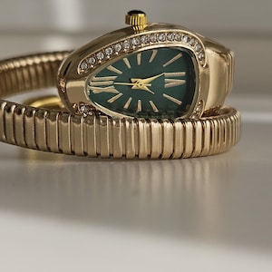 Może przedstawiać: Złoty zegarek na rękę z zieloną tarczą i diamentowymi akcentami. Zegarek ma unikalny owalny kształt i jest osadzony na elastycznej, złotej bransolecie. Tarcza zegarka posiada złote wskazówki i znaczniki.