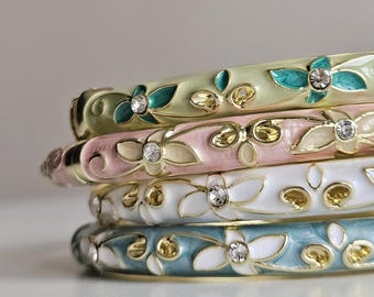 Könnte beinhalten: Ein Stapel von vier Emaille-Armreifen in Pastellfarben: Hellgrün, Rosa, Weiß und Blau. Jeder Armreif hat ein Blumenmuster mit goldenen Akzenten und funkelnden Kristallverzierungen. Die Armreifen sind übereinander gestapelt.