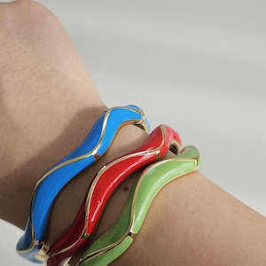 May include: Three colorful enamel bangles in blue, red, and green, with gold-colored trim. The wavy design adds a modern touch to these fashion accessories. These bracelets are a vibrant addition to any jewelry collection.