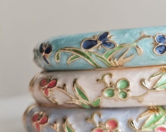 May include: Three stacked enamel bangles in shades of blue, pink, and white. Each bangle features a floral design with gold-colored accents. The top bangle is light blue with dark blue flowers. The middle bangle is white with pink and green flowers.