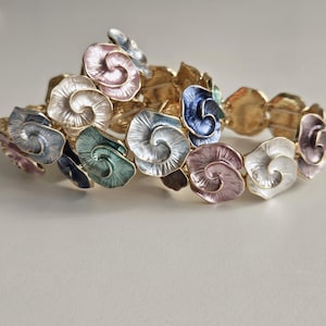 Op de afbeelding: Twee goudkleurige armbanden met kleurrijke emaille bloemmotieven. De bloemen zijn in tinten roze, blauw, groen en wit. De armbanden liggen op een witte ondergrond.