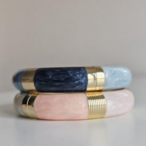 May include: Two stacked bangles. The top bangle has a navy blue and light blue marbled design with gold-colored metal accents. The bottom bangle has a pink and gray marbled design with gold-colored metal accents. The bangles are made of a hard material.