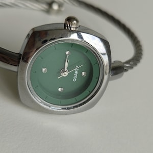 May include: A silver-toned quartz watch with a green face and a twisted metal band. The watch face is square with three small crystals and the word "QUARTZ" printed on it. The watch is on a white surface.