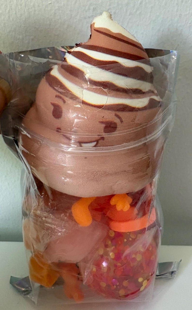 May include: A brown and white swirled plush toy shaped like a smiling character with a soft texture, packaged inside a clear plastic bag along with several small, colorful, translucent orange and pink plastic toy pieces and glittery spheres.