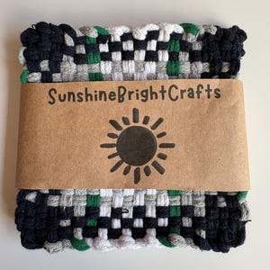 May include: A woven square in navy, green, white, and gray, with a brown paper band that reads "SunshineBrightCrafts" and features a sun graphic. The woven item is likely a trivet or hot pad.