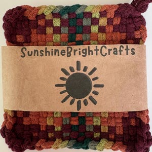 May include: A square woven potholder featuring a multicolored checkered pattern in shades of deep burgundy, orange, yellow, and dark green. A tan paper band wraps around the center, printed with the text SunshineBrightCrafts and a sun icon.