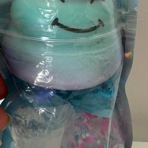 May include: A squishy toy shaped like a smiling creature with a pink top, teal middle, and purple base, featuring a white horn. It is packaged in a clear, iridescent plastic pouch along with a small, clear, round container filled with colorful confetti.