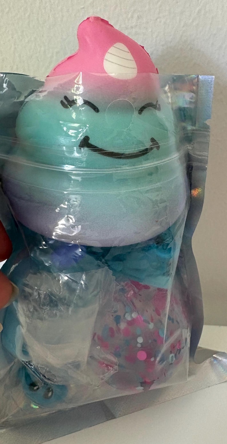 May include: A squishy toy shaped like a smiling creature with a pink top, teal middle, and purple base, featuring a white horn. It is packaged in a clear, iridescent plastic pouch along with a small, clear, round container filled with colorful confetti.