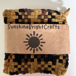 May include: Woven square pot holder in shades of black, brown, and gold. A brown paper band with the text "SunshineBrightCrafts" and a sun graphic is wrapped around the pot holder. The pot holder is made of woven fabric.
