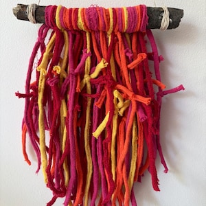 May include: A wall hanging featuring a dark brown wooden branch wrapped with strips of fabric in shades of hot pink, orange, and yellow. Numerous long, knotted fabric strips in the same vibrant color palette hang downward from the branch.