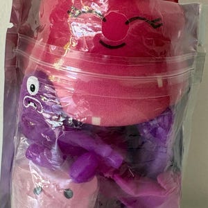 May include: A clear plastic pouch containing a plush strawberry toy with a green top and a smiling face, a purple plastic octopus figure, a small light pink container with a face, and a clear sphere filled with purple glittery confetti pieces.