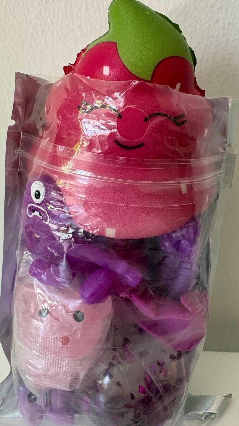 May include: A clear plastic pouch containing a plush strawberry toy with a green top and a smiling face, a purple plastic octopus figure, a small light pink container with a face, and a clear sphere filled with purple glittery confetti pieces.