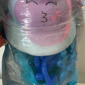 May include: A clear plastic pouch containing a plush toy with a purple, pink, and light blue face featuring a smiling expression with closed eyes. Below the plush, the pouch holds blue plastic components and a base filled with colorful glitter.