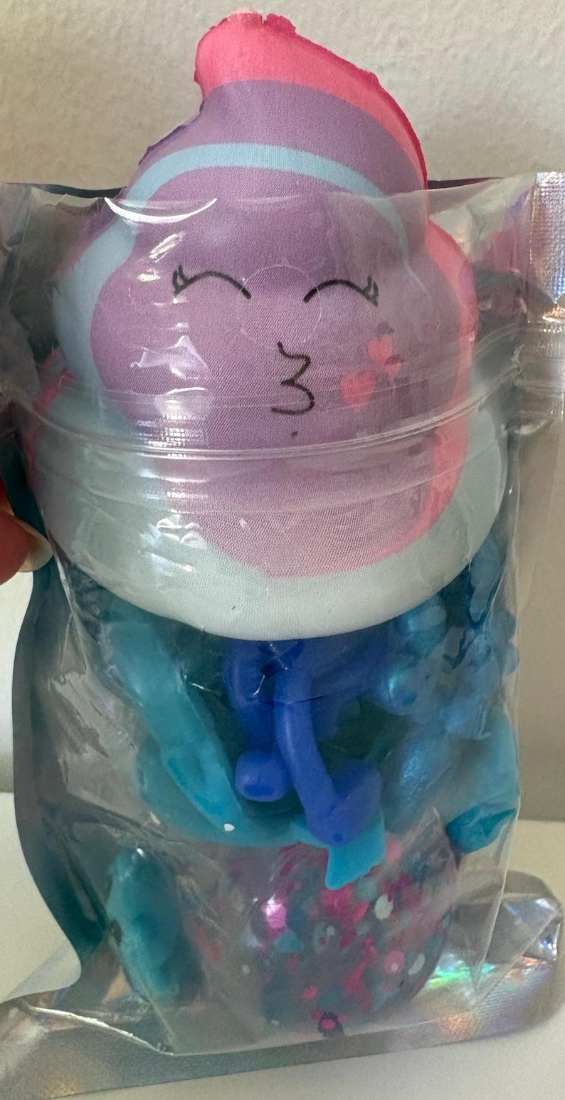 May include: A clear plastic pouch containing a plush toy with a purple, pink, and light blue face featuring a smiling expression with closed eyes. Below the plush, the pouch holds blue plastic components and a base filled with colorful glitter.