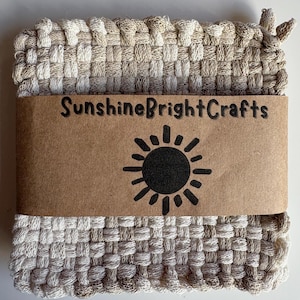 May include: A square, woven trivet in shades of beige and cream. A brown paper band wraps around the trivet, displaying the text "SunshineBrightCrafts" and a black sun graphic. The trivet is made of a thick, textured material.