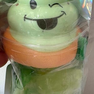 May include: A light green squishy toy shaped like a swirled ice cream cone with a brown topping and a winking face emoji. The toy features a smiling expression with a tongue sticking out and is enclosed in a clear, iridescent plastic packaging.