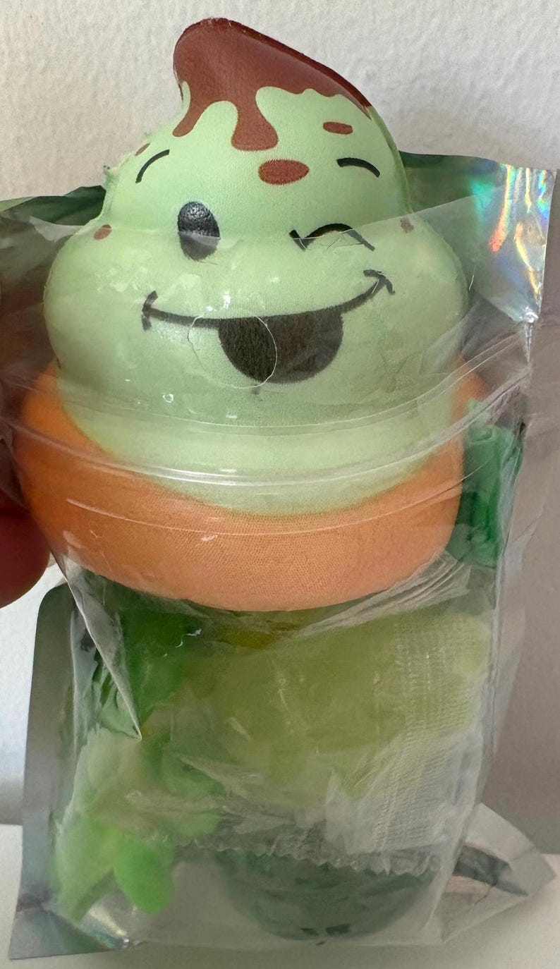 May include: A light green squishy toy shaped like a swirled ice cream cone with a brown topping and a winking face emoji. The toy features a smiling expression with a tongue sticking out and is enclosed in a clear, iridescent plastic packaging.