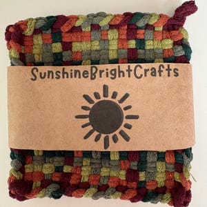 May include: A woven square hot pad in shades of red, green, orange, and olive. The hot pad has a loop for hanging. A brown paper band with the text "SunshineBrightCrafts" and a sun graphic is wrapped around the pad.