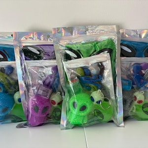 May include: Four iridescent, resealable plastic pouches containing alien-themed party favor sets. Each set includes a textured, spiky alien figure, a small alien keychain, and a miniature alien figurine, all packaged in clear, holographic bags.