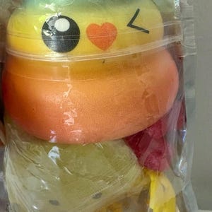 May include: A soft, squishy toy shaped like a coiled pile, featuring a vibrant rainbow gradient from blue at the top to yellow and orange. It has a printed face with large black eyes, a small orange heart, and a winking expression, all sealed in a clear plastic bag.