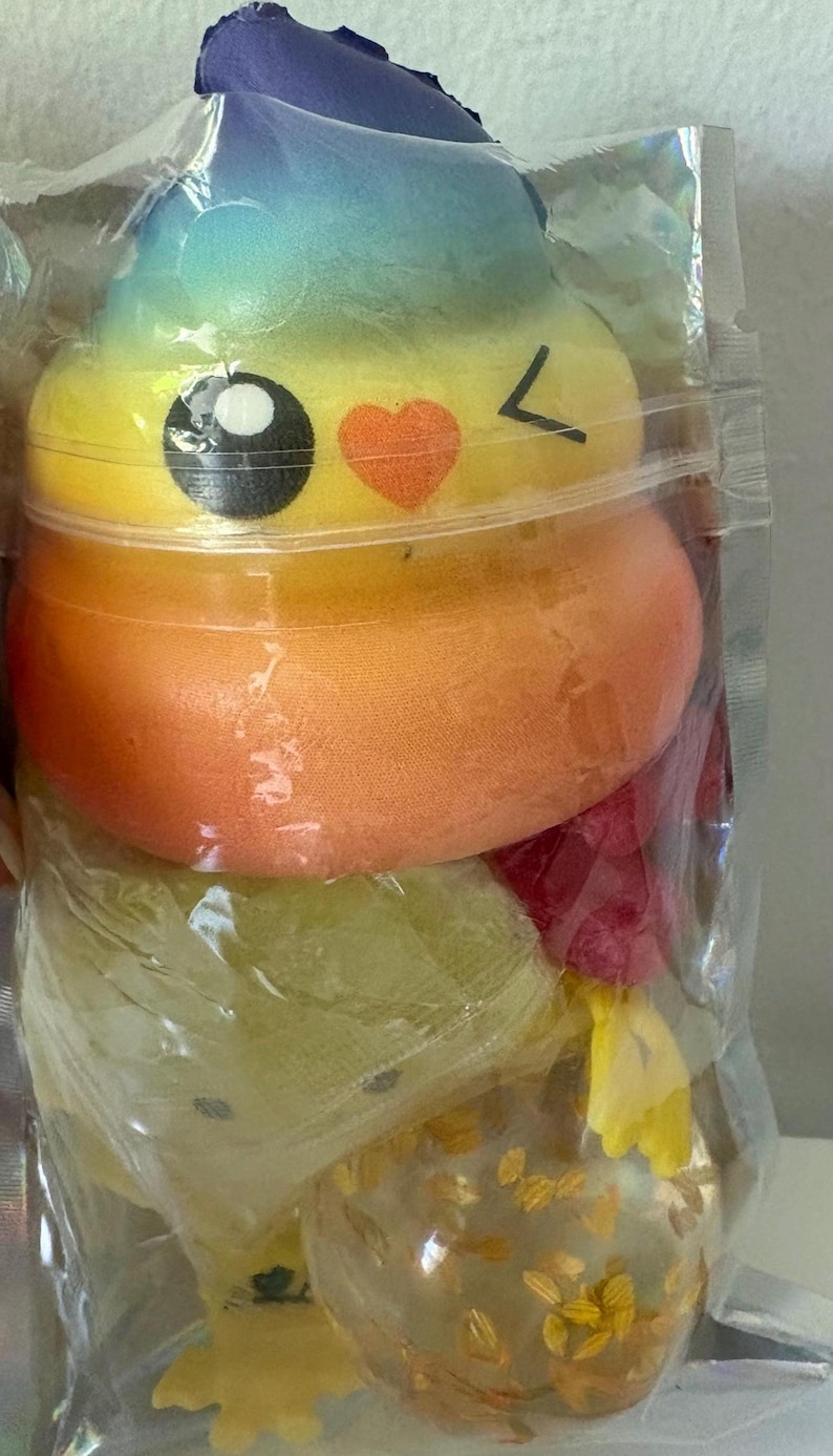 May include: A soft, squishy toy shaped like a coiled pile, featuring a vibrant rainbow gradient from blue at the top to yellow and orange. It has a printed face with large black eyes, a small orange heart, and a winking expression, all sealed in a clear plastic bag.