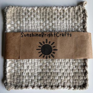May include: A square woven potholder featuring a checkered pattern in shades of beige, cream, and white. A brown paper band wraps around the center, printed with the text SunshineBrightCrafts and a black sun icon with radiating rays.