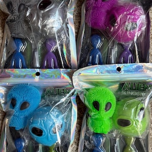 May include: Four packages of alien-themed toys. Each package contains a large alien head, a small alien figure, and a slingshot. The alien heads are gray, purple, blue, and green. The small figures are blue, purple, and green.
