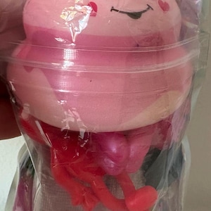 May include: A pink squishy toy shaped like a swirled dessert with a smiling face, a small bow, and heart-shaped cheeks. It is packaged in a clear plastic bag along with red plastic appendages and a small container filled with colorful confetti pieces.