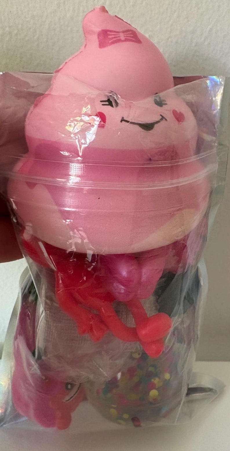 May include: A pink squishy toy shaped like a swirled dessert with a smiling face, a small bow, and heart-shaped cheeks. It is packaged in a clear plastic bag along with red plastic appendages and a small container filled with colorful confetti pieces.