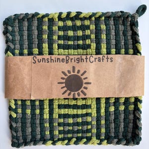 May include: A square woven potholder featuring a geometric pattern in dark green, lime green, and grey yarn. A brown paper band wraps around the center, printed with the text SunshineBrightCrafts and a dark brown sun icon with radiating rays.
