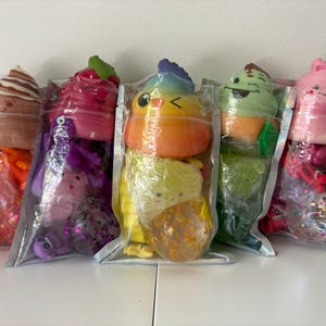 May include: Five clear plastic pouches each containing a colorful squishy toy shaped like a swirl of soft serve ice cream with a smiling face, accompanied by smaller assorted sensory toys including glittery balls and small animal figures.
