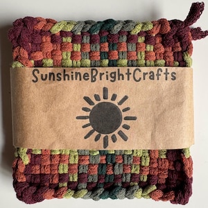 May include: A woven square hot pad in shades of burgundy, orange, green, and gray. A brown paper band wraps around the pad, displaying the text "SunshineBrightCrafts" and a sun graphic. The hot pad has a loop for hanging.