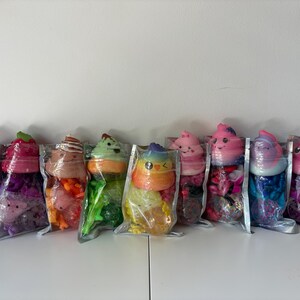 May include: Eight clear plastic pouches containing colorful squishy toys. Each pouch features a soft, shaped topper resembling a swirl or character with a smiling face, sitting atop a base filled with various small, vibrant plastic charms and beads.