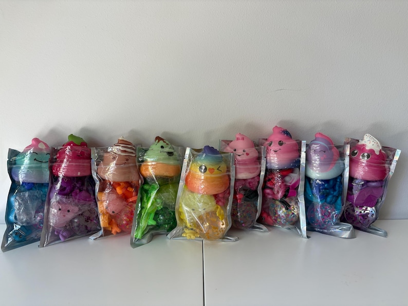 May include: Eight clear plastic pouches containing colorful squishy toys. Each pouch features a soft, shaped topper resembling a swirl or character with a smiling face, sitting atop a base filled with various small, vibrant plastic charms and beads.