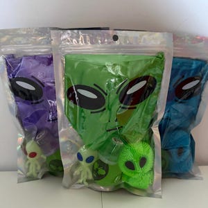 May include: Three clear, resealable pouches featuring a printed alien face graphic. Each pouch contains a small alien figurine, a round container with an alien face, and a textured, spiky alien-shaped toy in purple, green, or blue colors.