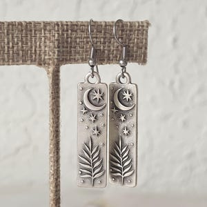 May include: A pair of silver-toned rectangular drop earrings featuring an embossed design of a crescent moon, multiple stars, and a fern-like leaf. The earrings hang from simple metal hooks and have a matte, oxidized finish.