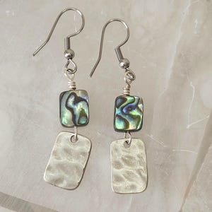 May include: A pair of dangle earrings with silver-tone hooks. Each earring features a square, iridescent abalone bead above a hammered, rectangular silver-tone charm. The earrings are displayed against a white background.