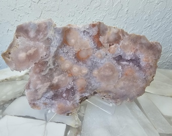 May include: A large, irregularly shaped, polished agate specimen. The stone features a mix of colors, including lavender, peach, and white, with a crystalline structure. The agate is displayed on a clear stand, showcasing its natural beauty.