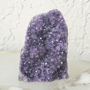 May include: A large, raw amethyst crystal cluster. The purple crystal has a rough, textured surface with many small, sparkling crystal points. The amethyst sits on a white surface against a textured white background.